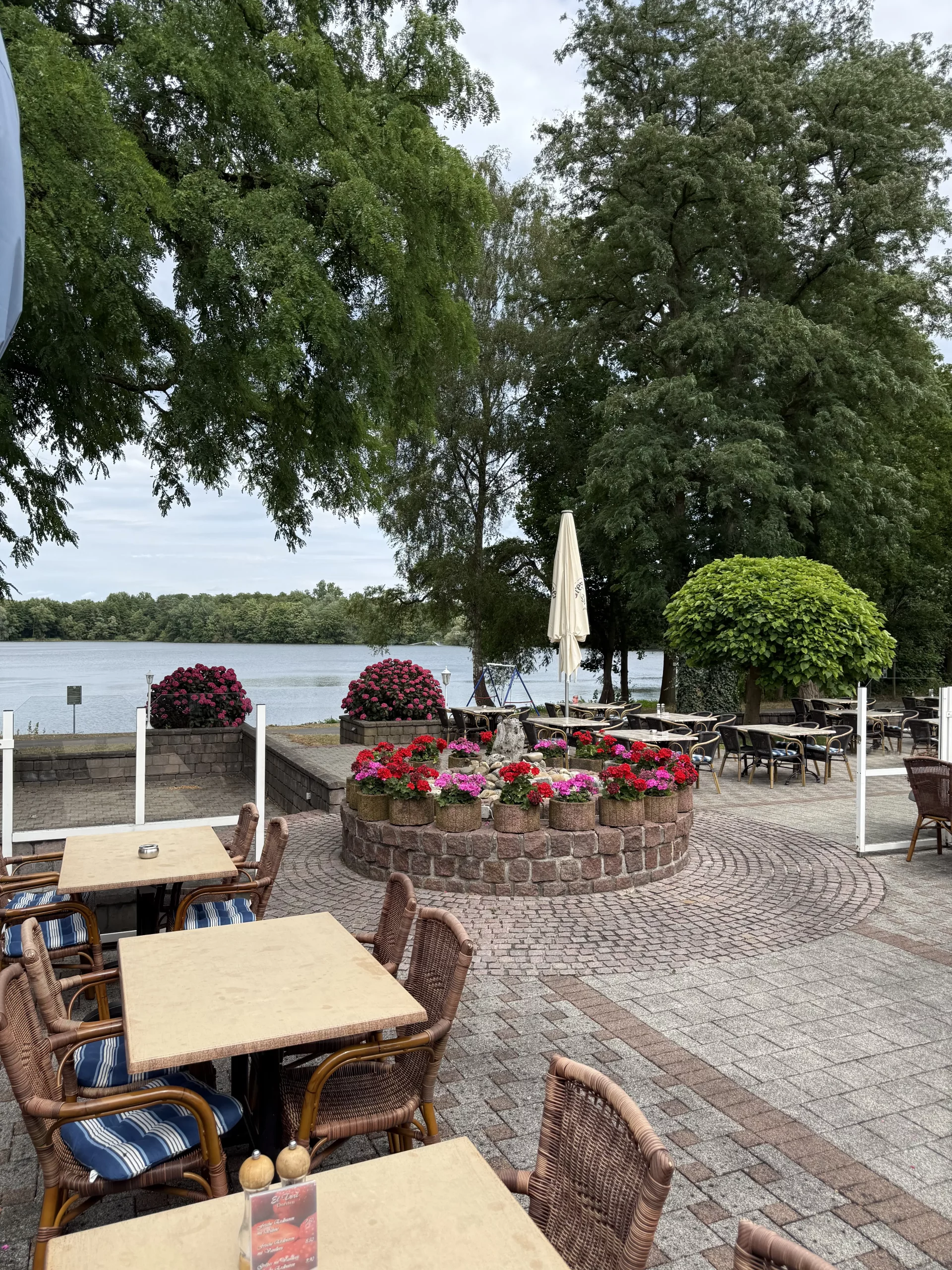 Caféterrasse am See mit Blumenbeeten und Bäumen.