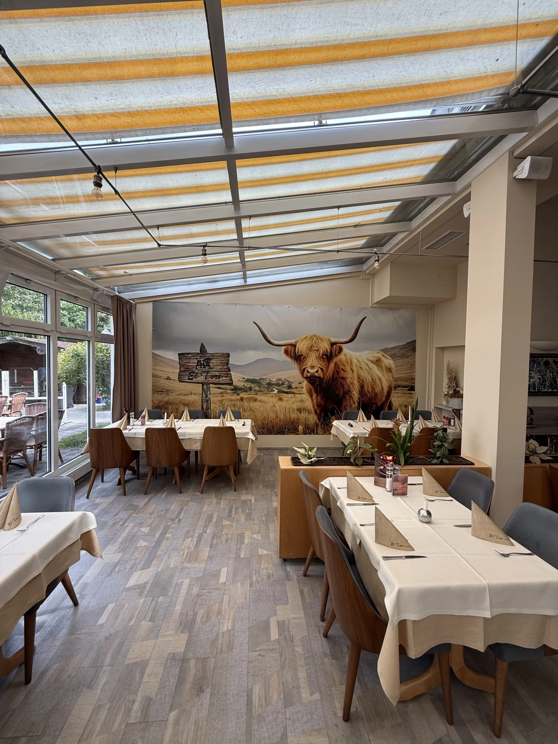 Helles Restaurant mit Glasdach, Kuh-Wandbild und Tischen